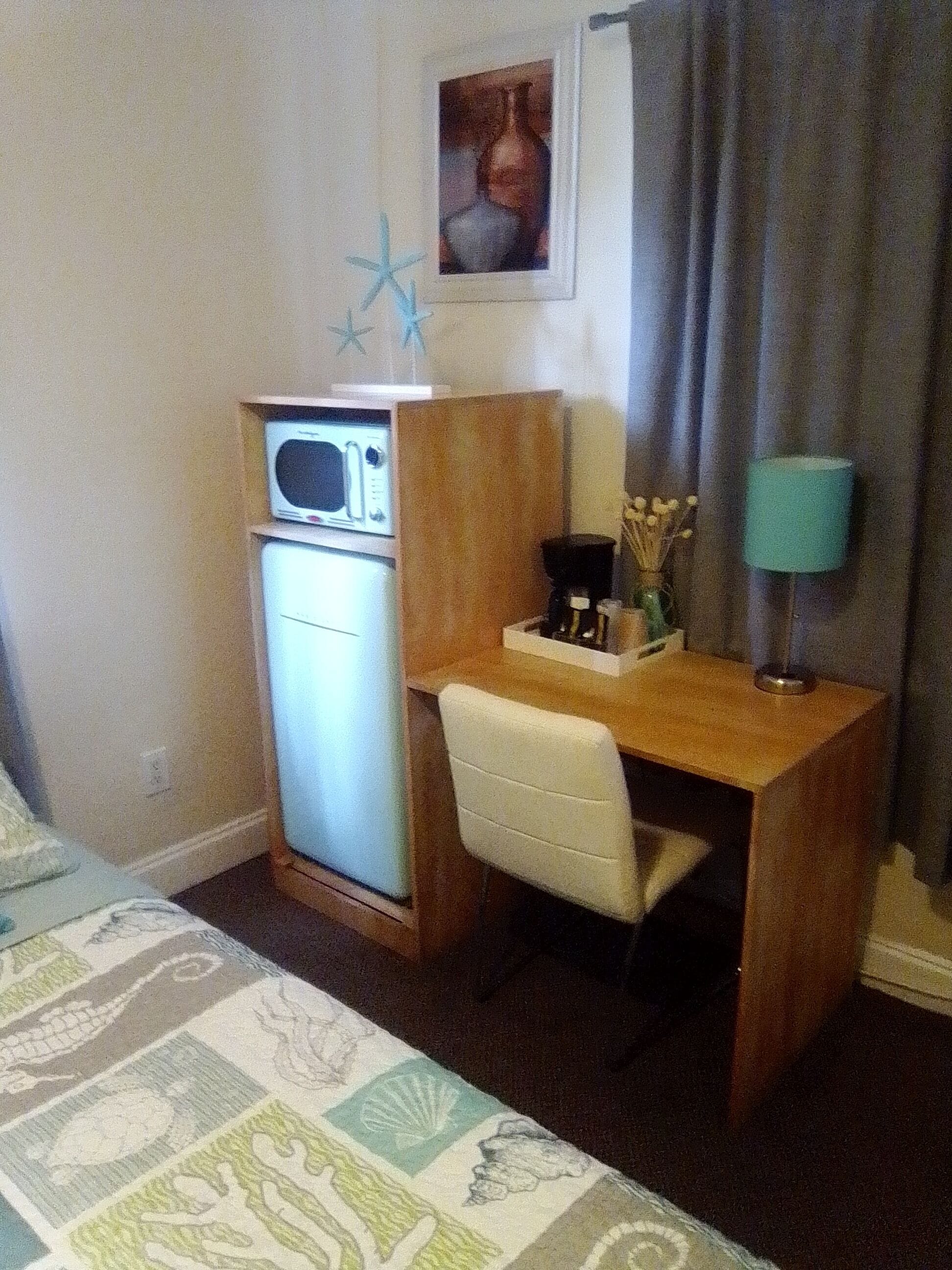 A compact hotel room with a kitchenette, a small desk, a chair, a bed, and a single window with curtains.