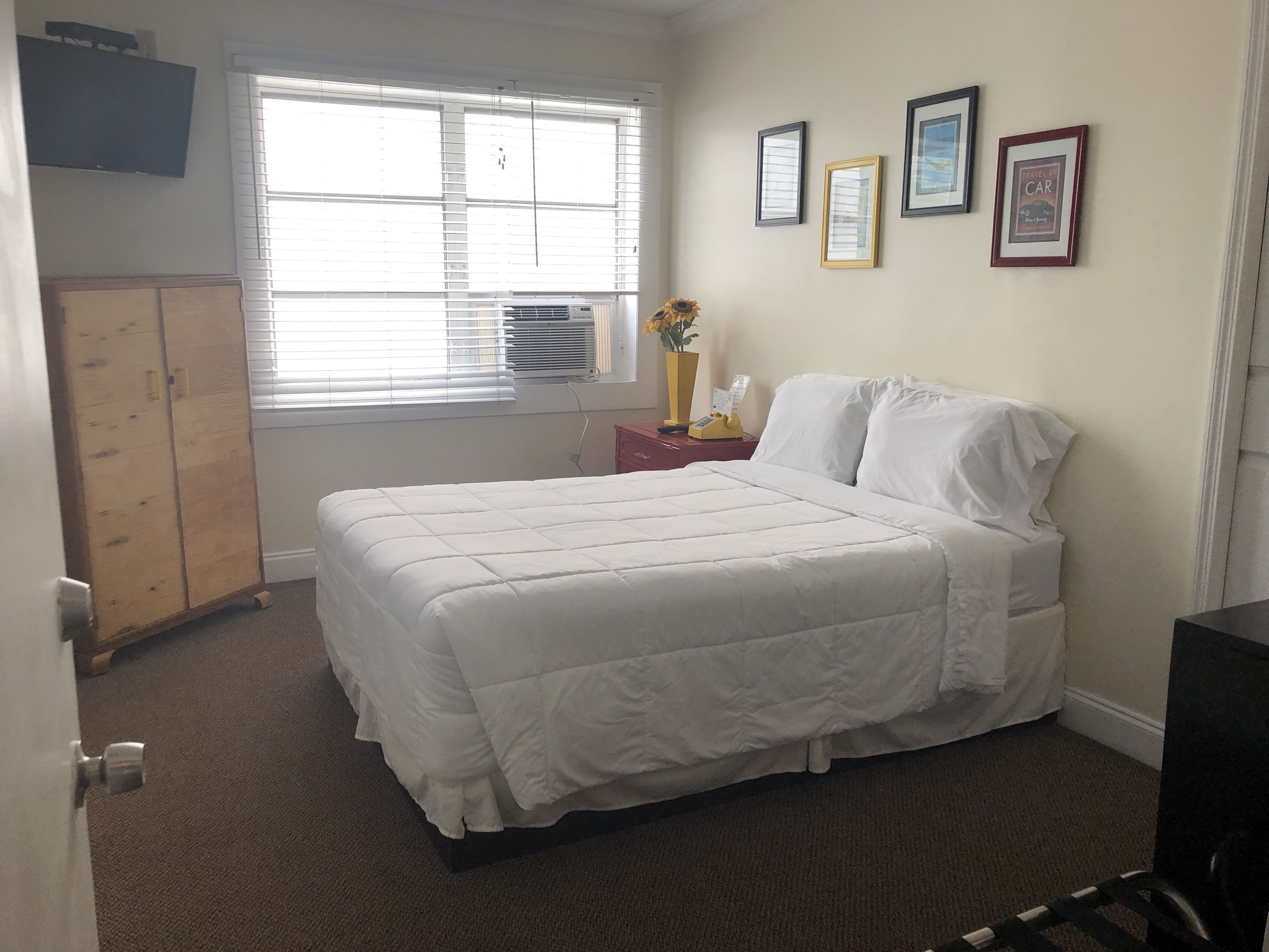 Une chambre à coucher avec un lit, une armoire en bois, une fenêtre avec des stores et des tableaux encadrés accrochés au mur.