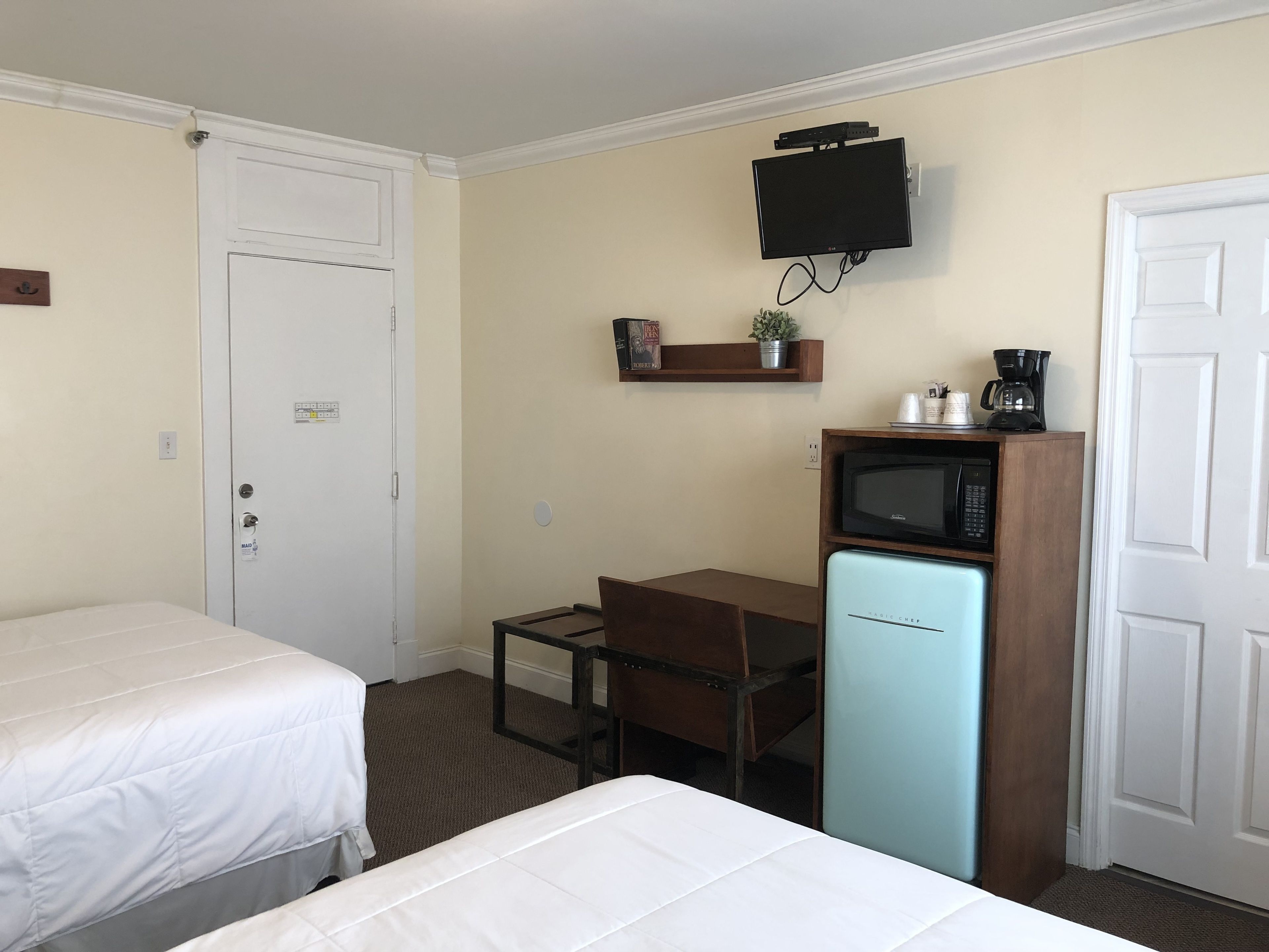 Habitación de hotel con dos camas individuales, un escritorio pequeño con una silla, microondas, televisor montado en la pared y cafetera en un estante.