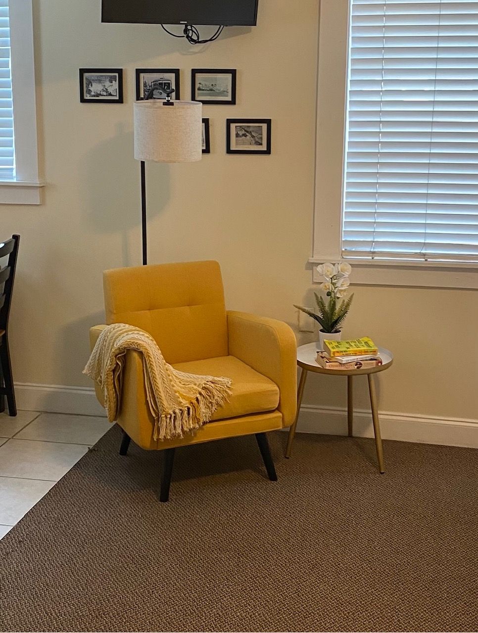 Uma poltrona amarela com um cobertor, uma luminária de chão, uma mesinha com livros e uma planta, e quadros nas paredes.