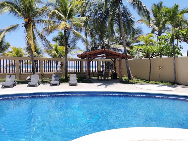 Piscina con sillas de descanso y una pérgola de madera, rodeada de palmeras y vista a la playa.
