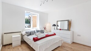 A modern bedroom with a large bed, a window, a bedside table with a lamp, and a dresser with a mirror. - 2 Bed Flat, Dedicated Workspace, Parking, 4KTV (London)