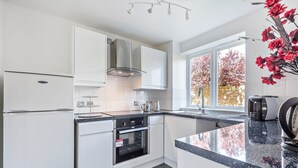 A modern kitchen with white cabinetry, a stainless steel range hood, a built-in oven, a large window with autumn foliage view, and a coffee maker on the counter. - 2 Bed Flat, Dedicated Workspace, Parking, 4KTV (London)