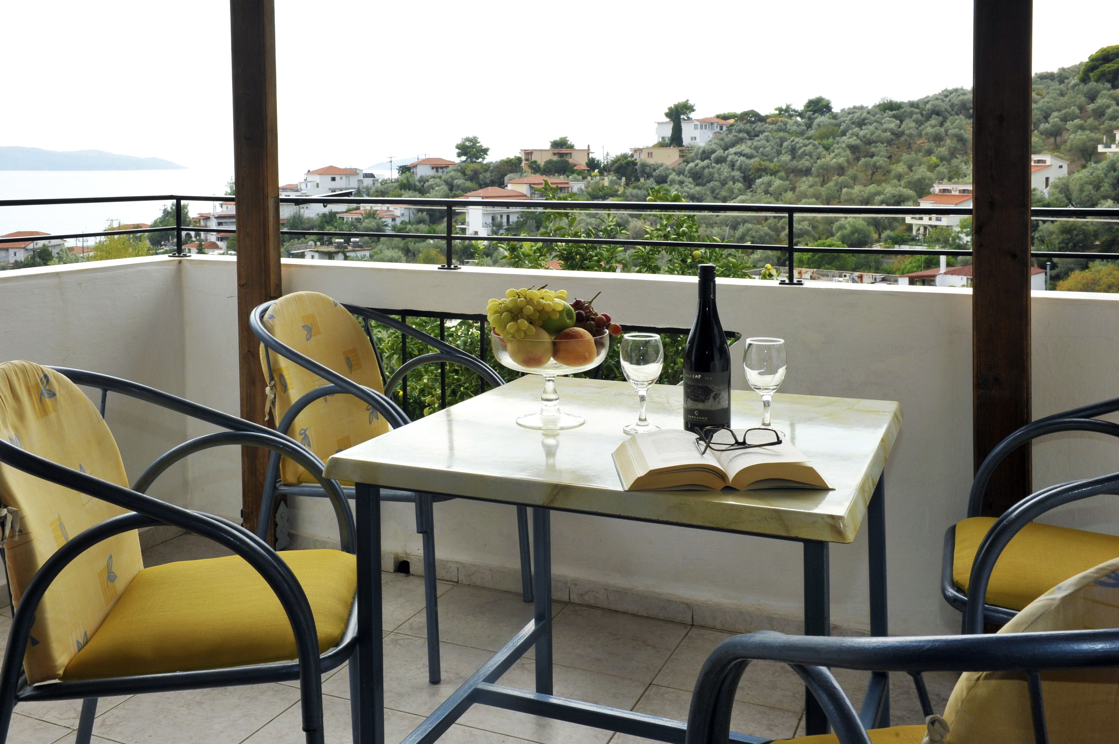 A balcony with yellow chairs, a table with a book, a bottle, and a glass of wine.