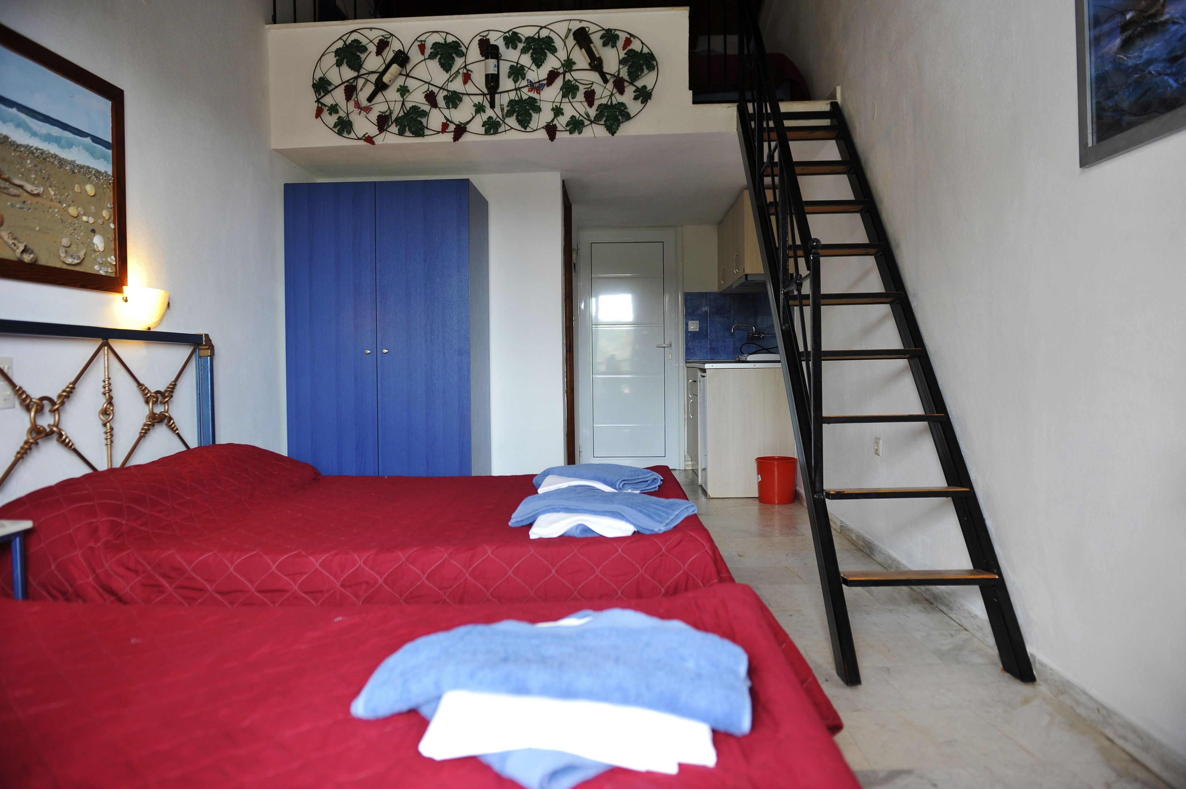 Une chambre avec deux lits, une armoire bleue, une échelle et, au fond, un coin cuisine.