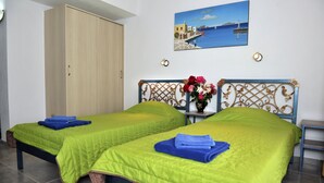 A room with two beds, each with a green bedspread and blue towels, a wooden wardrobe, a wall-mounted painting, and a small table with a vase of flowers. - Fengeros Village (Skiathos)