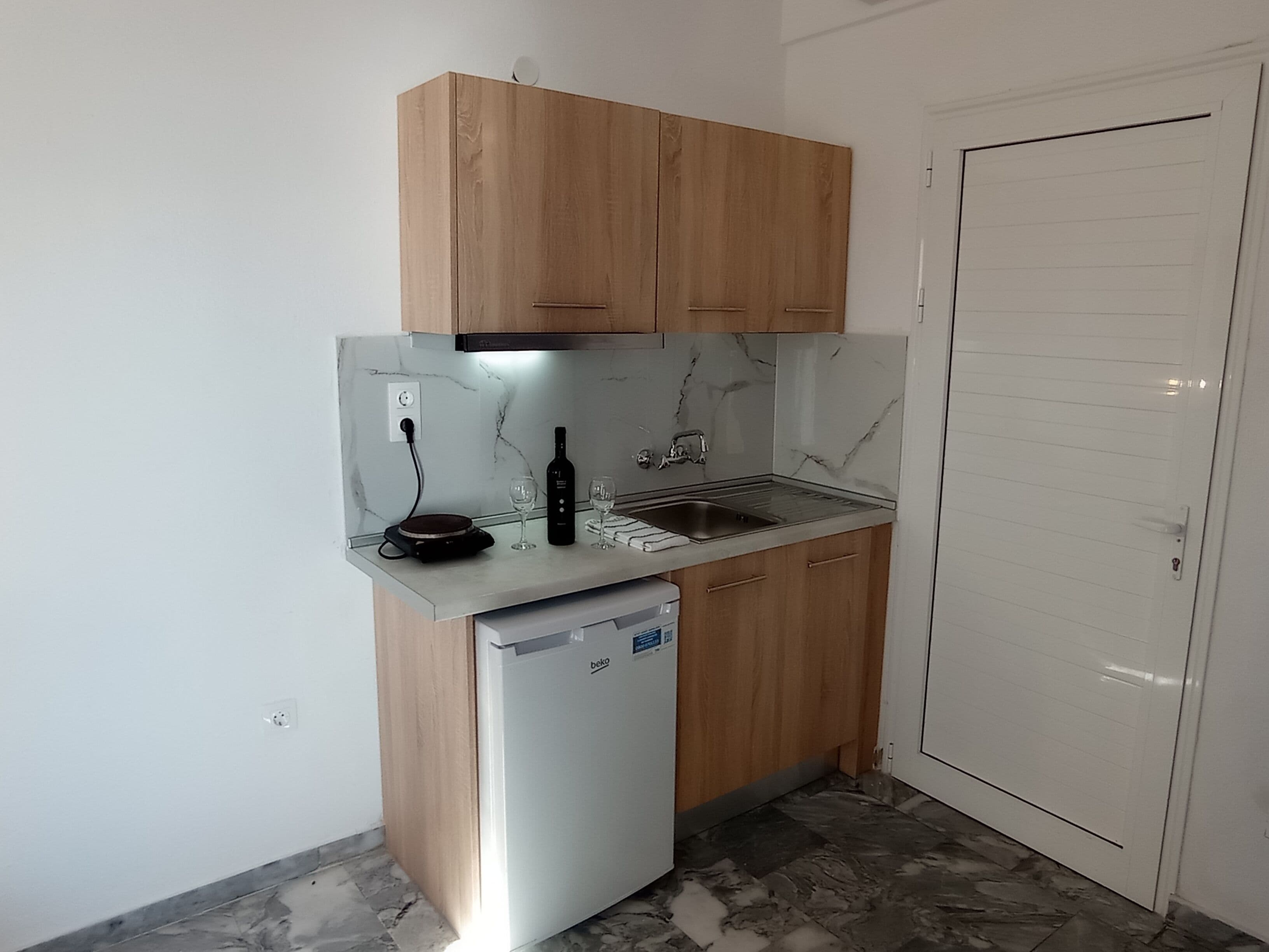 A compact kitchen with wooden cabinets, a built-in dishwasher, a marble backsplash, and a white door.