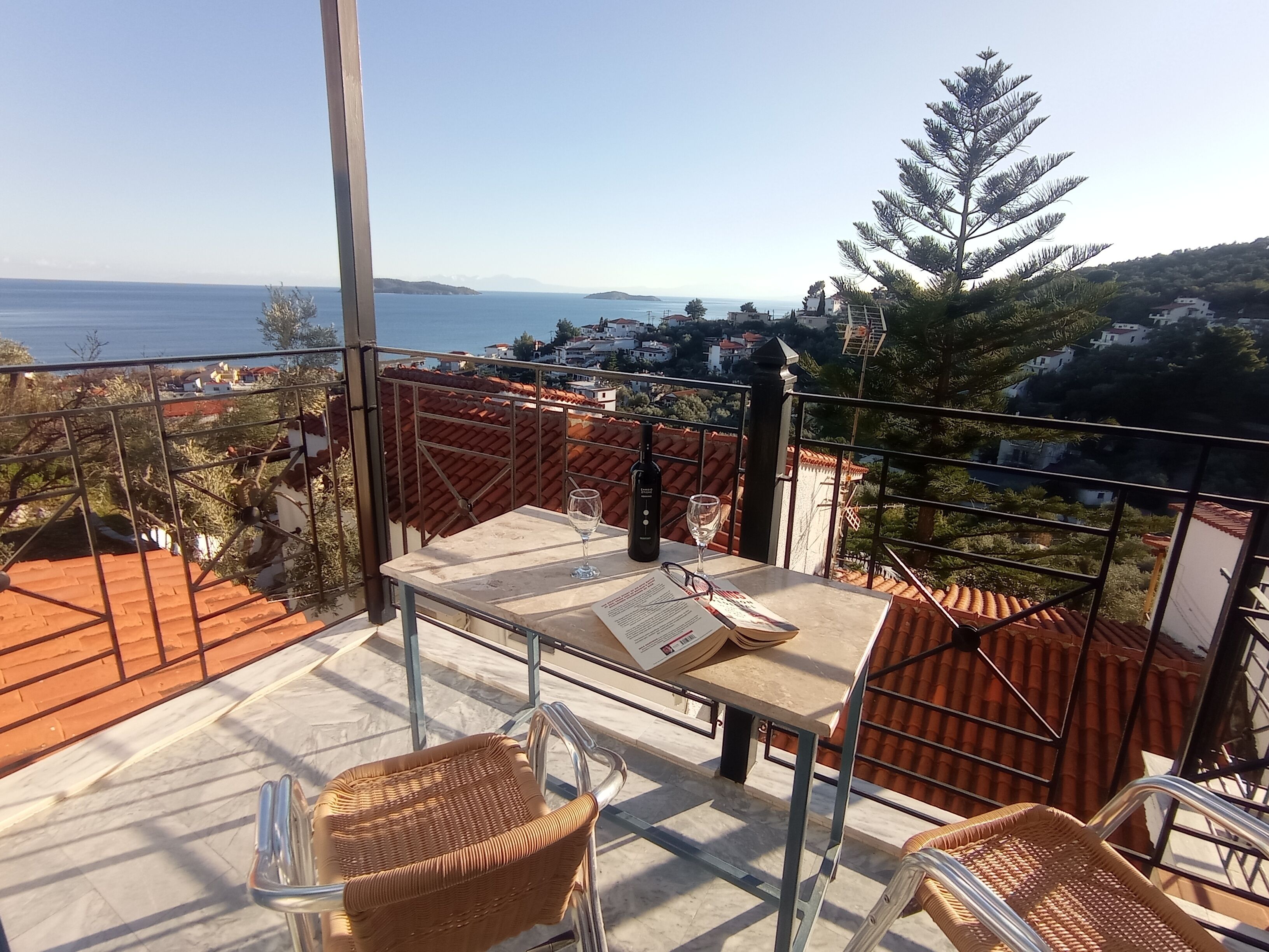 Una terraza con mesa, sillas y una botella de vino, con vistas a un pueblo costero de edificios con tejados rojos y cielo despejado.