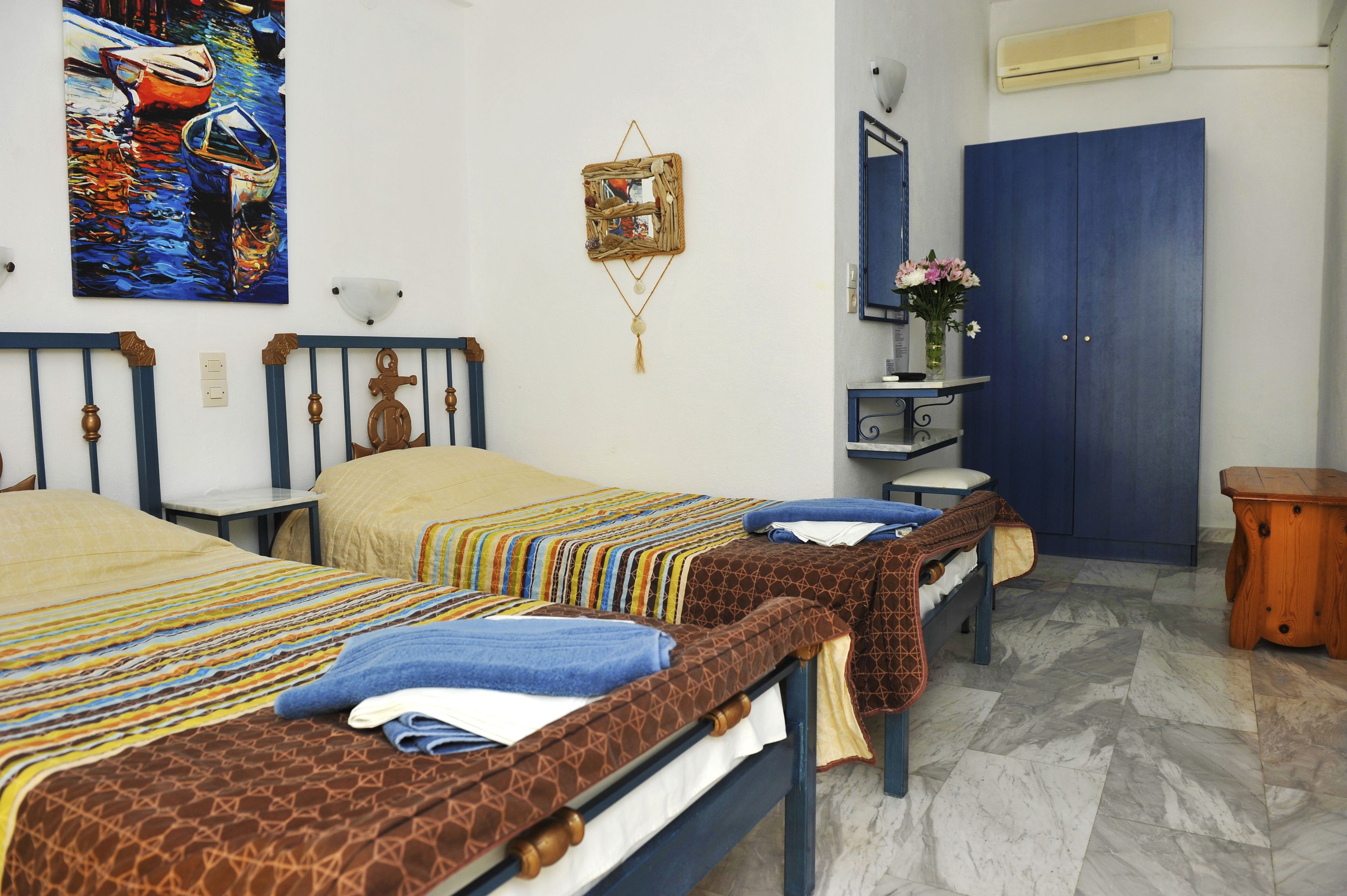A hotel room with two beds, a blue wardrobe, a small table with a vase of flowers, and a painting on the wall.