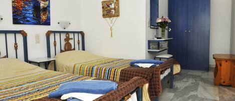 A hotel room with two beds, a blue wardrobe, a small table with a vase of flowers, and a painting on the wall.