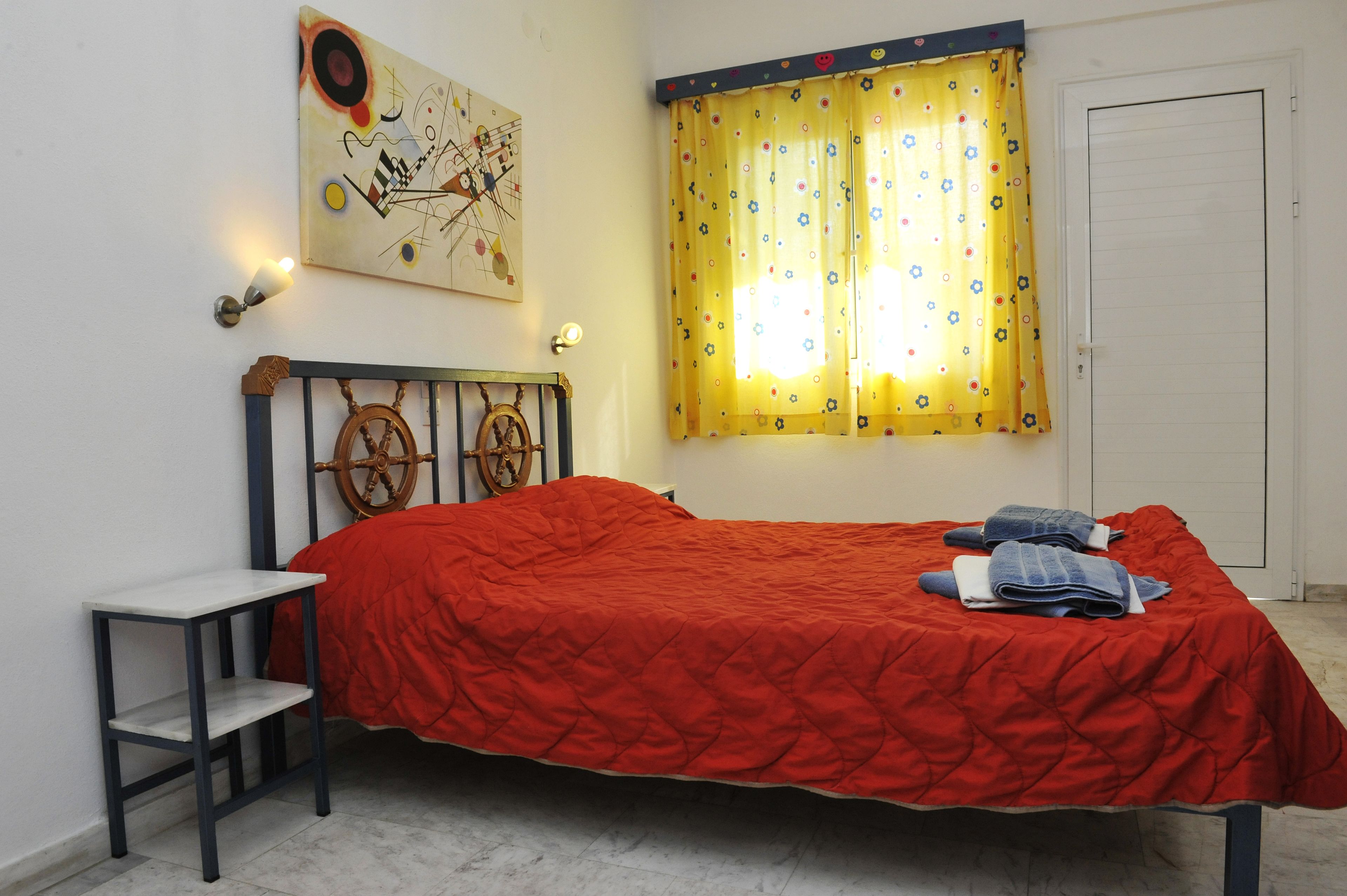 Ein Schlafzimmer mit einem Bett, einem kleinen Tisch, einer Wandleuchte, einer Tür und einem Fenster mit Vorhang.