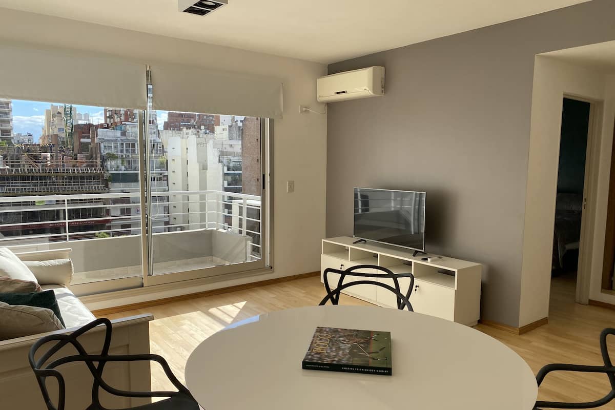 A modern living room with a balcony, a flat-screen TV, a round dining table, and a sofa.
