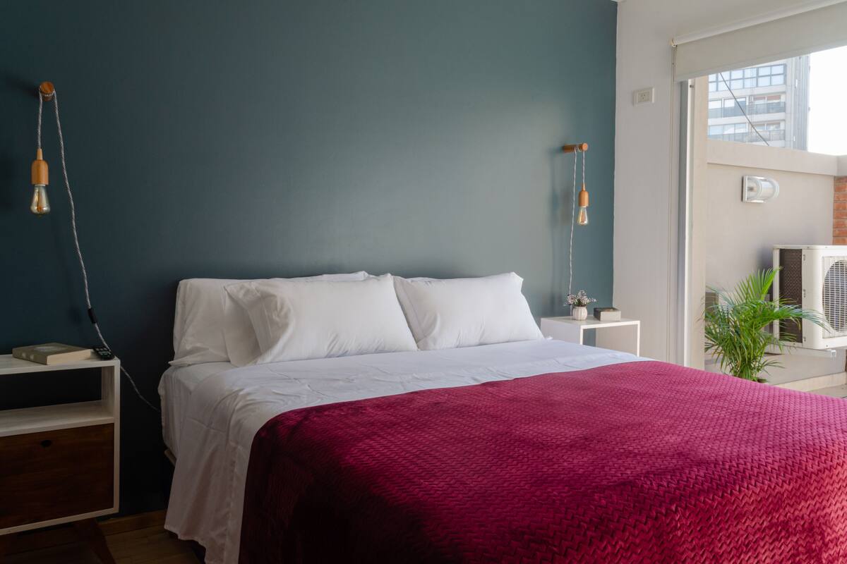 A bedroom with a bed, white bedding, a red blanket, a nightstand, a lamp, and a potted plant.