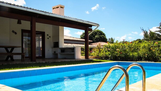 A swimming pool with a covered patio area, a bench, and a view of a house with a tiled roof. - Pipa Casa Eduardo (Tibau do Sul)