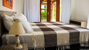 A bedroom with a checkered bedspread, a bedside lamp, and a view of a garden through a door. - Pipa Casa Eduardo (Tibau do Sul)