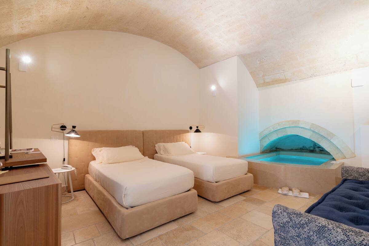A hotel room with two beds, a small table, a desk, a lamp, and a bathtub with a stone arch.