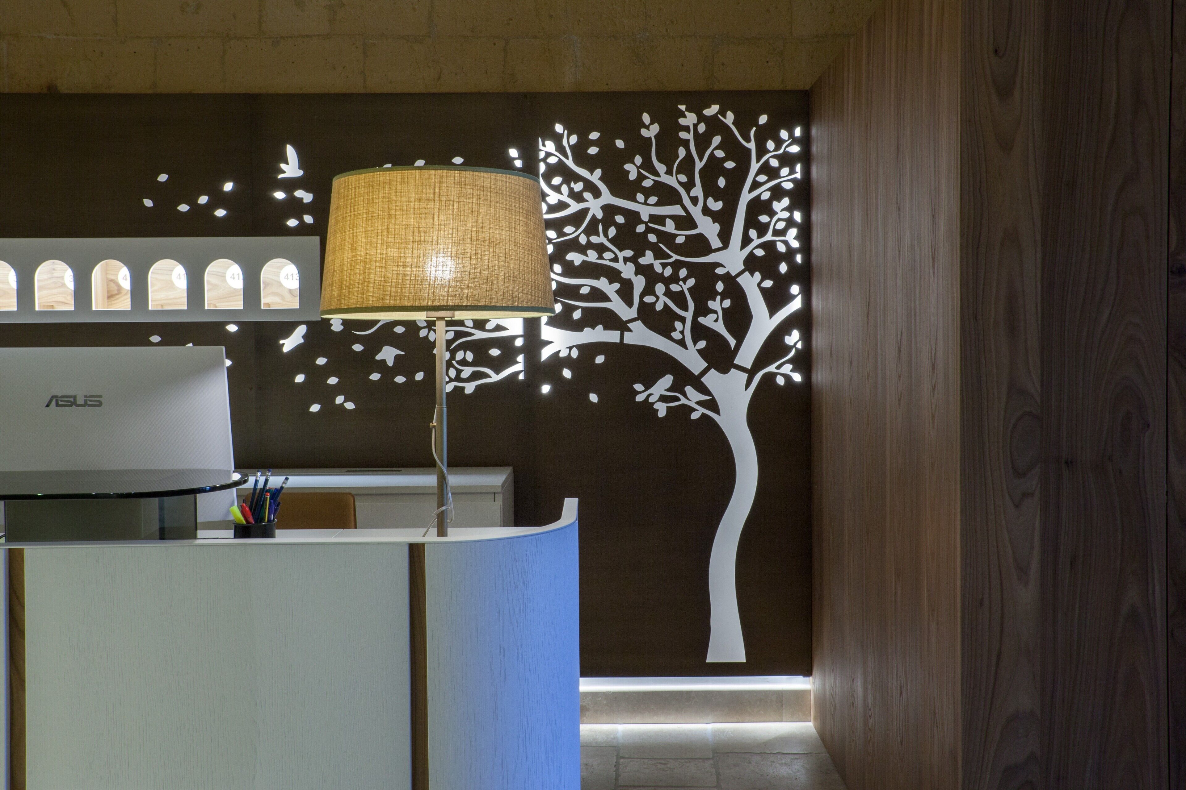 A reception desk with a modern lamp, a wall-mounted tree decoration, and a series of arched windows.