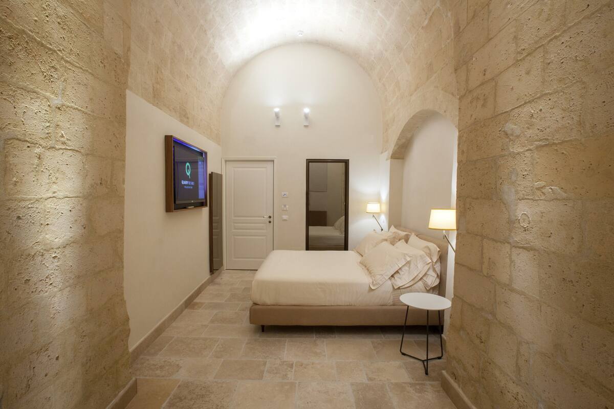A narrow hallway leading to a bedroom with a bed, a television, a door, and a small table with a lamp.