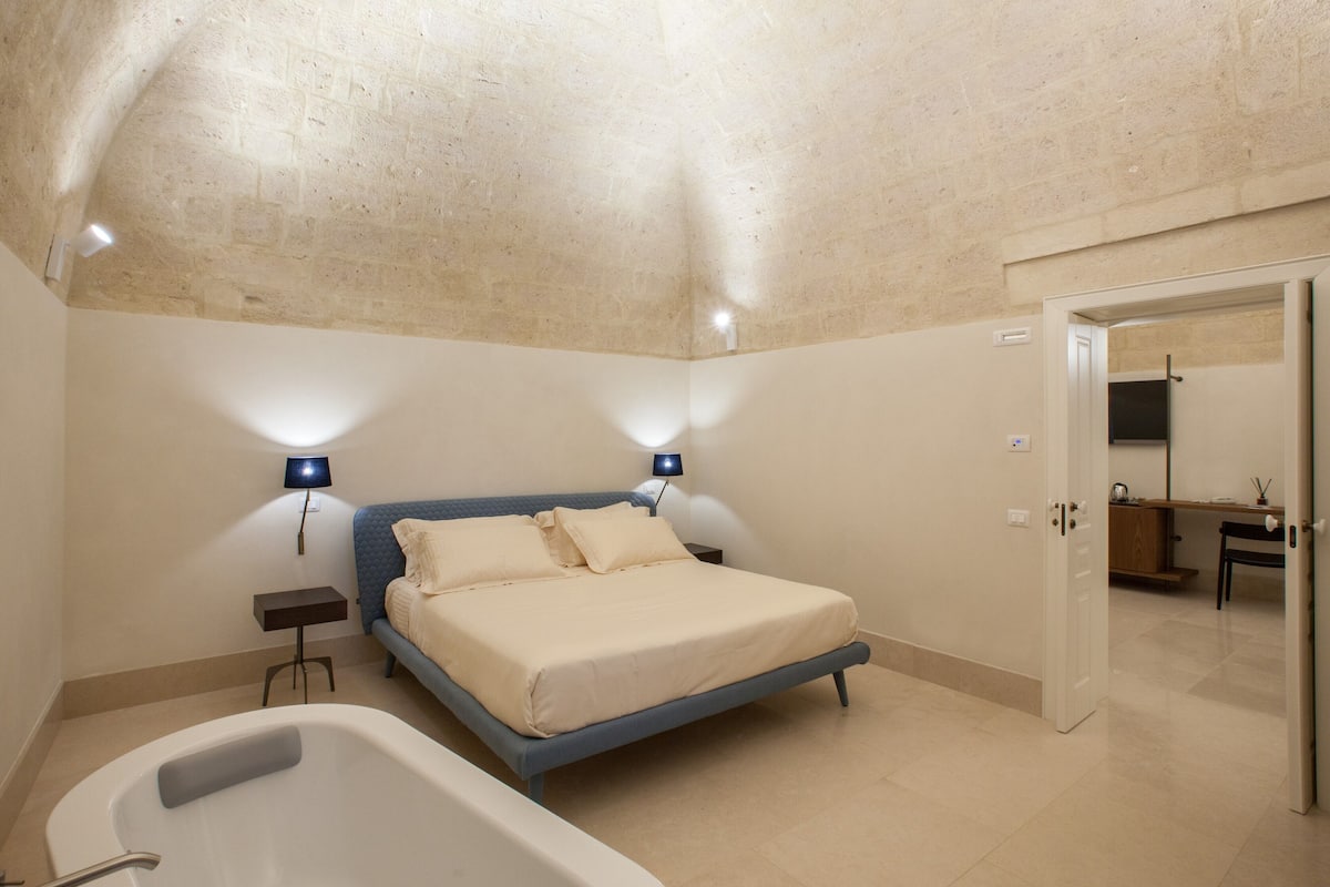 A bedroom with a bed, bedside lamps, a nightstand, a bathtub, and a door leading to another room.