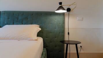 Una cama bien hecha con cabecera tapizada de verde, una mesita redonda con lámpara y un enchufe en la pared.