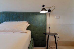 Una cama bien hecha con cabecera tapizada de verde, una mesita redonda con lámpara y un enchufe en la pared.