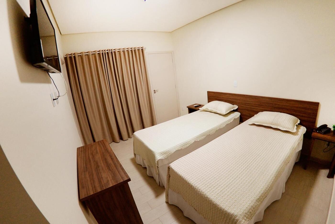 Quarto de hotel com duas camas de solteiro, uma escrivaninha de madeira, televisão fixada na parede e cortina.