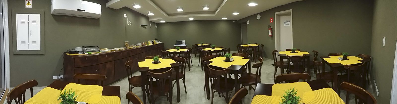 Uma área de refeições com várias mesas e cadeiras, um balcão com um computador e papéis, e um extintor de incêndio na parede.