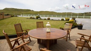 A round wooden table with chairs and a lantern, set on a paved patio with a white fence and flags in the background. - The 19th Lodge Ballybunion (Ballybunion)