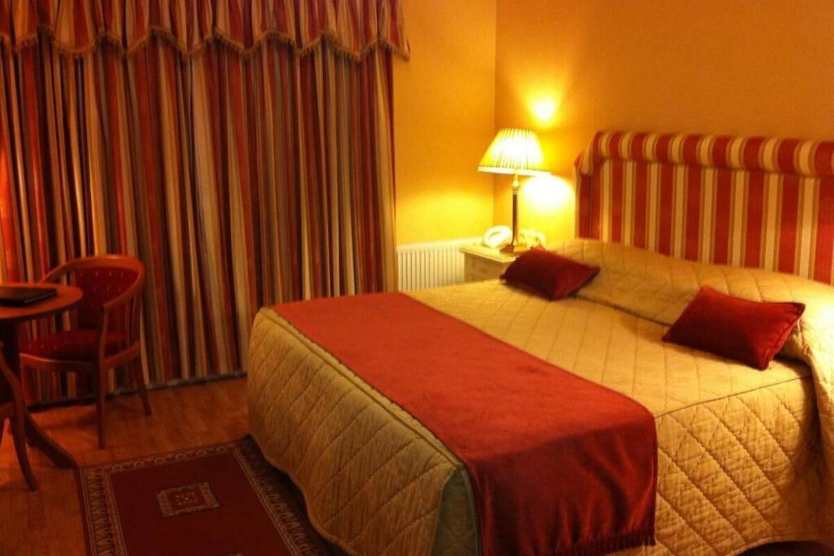 A hotel room with a large bed, a bedside table with a lamp, a chair, and a window with curtains.