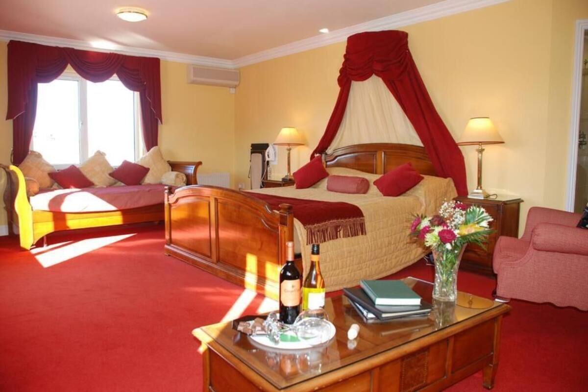 A hotel room with a large bed, a bedside table with a wine bottle and glasses, a chair, and a desk with a vase of flowers.
