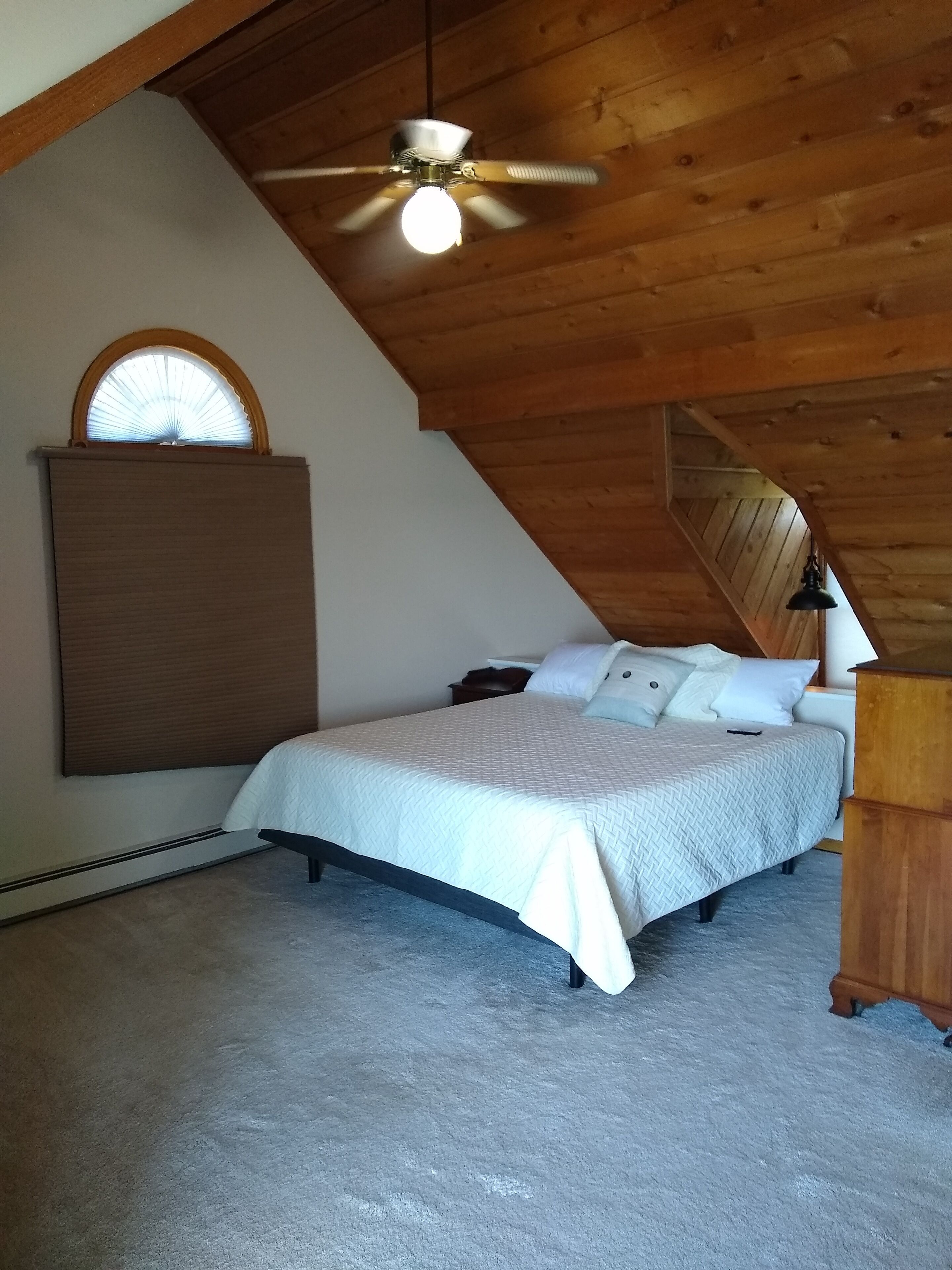 A bedroom with a sloped ceiling, a bed with white bedding, a wooden headboard, a nightstand, a desk, and a ceiling fan.