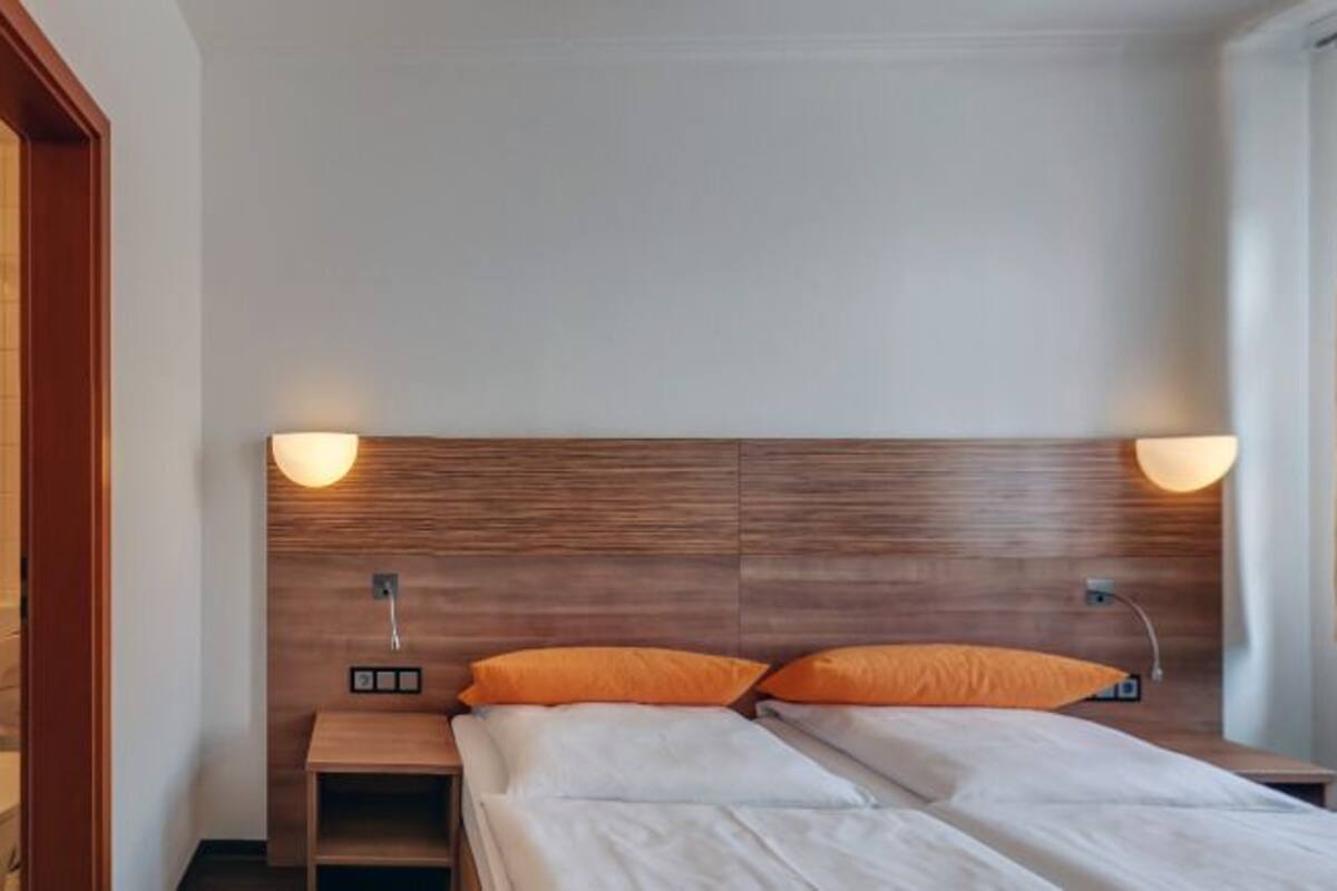 Une chambre d’hôtel avec une tête de lit en bois, deux oreillers, une table de chevet et une fenêtre avec des rideaux.