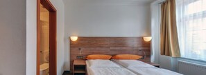 A hotel room with a wooden headboard, two pillows, a bedside table, and a window with curtains. - Hotel Drei Kaiserberge (Goeppingen)