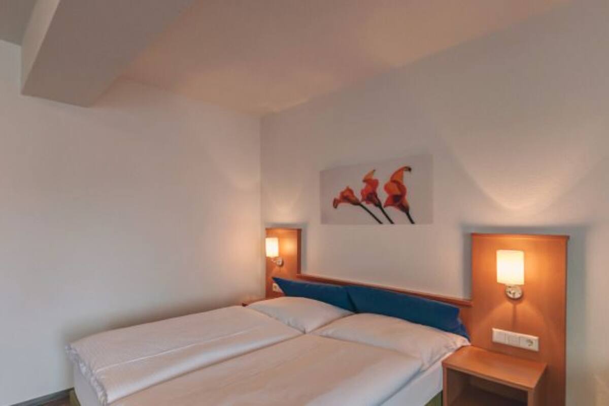 Une chambre d’hôtel avec un lit, une porte en bois, une lampe fixée au mur et une petite table.