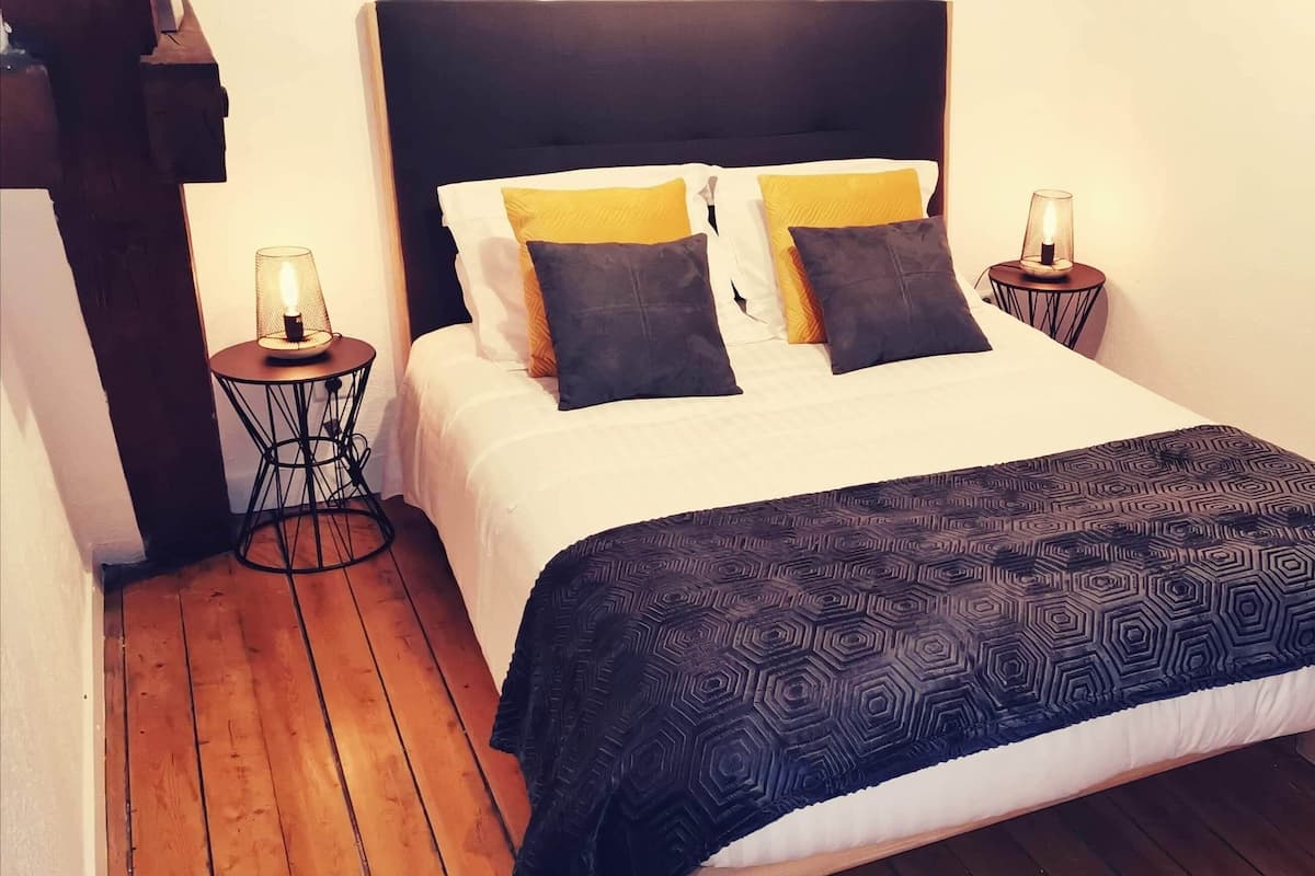 Un lit bien fait, recouvert d’une couverture à motifs sombres, deux tables de chevet avec des lampes et un plancher en bois.