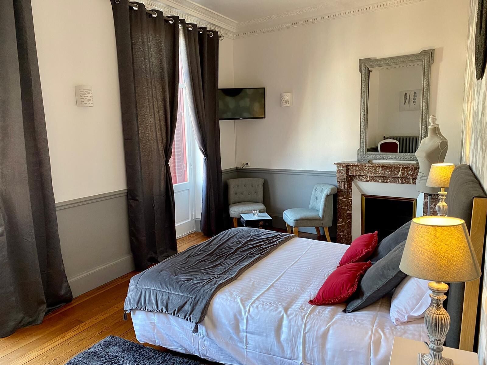 A hotel room with a bed, a chair, a desk, a mirror, and a fireplace.