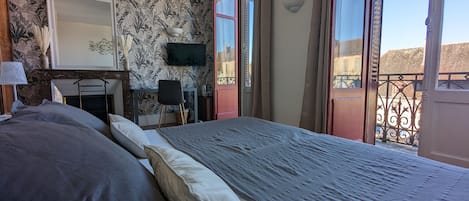 A bedroom with a large bed, a desk, a chair, a TV, and a balcony with a wrought iron railing.