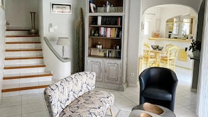 Un salon avec un canapé à motifs, une bibliothèque, un coin repas avec des chaises jaunes et un escalier.