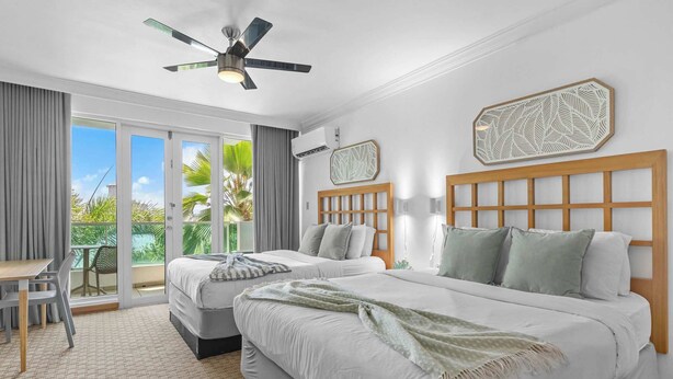 A hotel room with two beds, a ceiling fan, a balcony with a table and chairs, and a view of palm trees. - Island Studio | Stunning Waterfront Getaway (Condado Lagoon Villas)