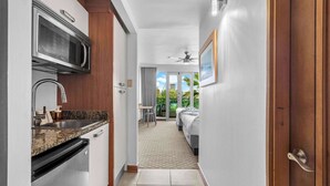 A narrow hallway with a kitchen on the left, a bed with a view of greenery on the right, and a ceiling fan in the center. - Island Studio | Stunning Waterfront Getaway (Condado Lagoon Villas)