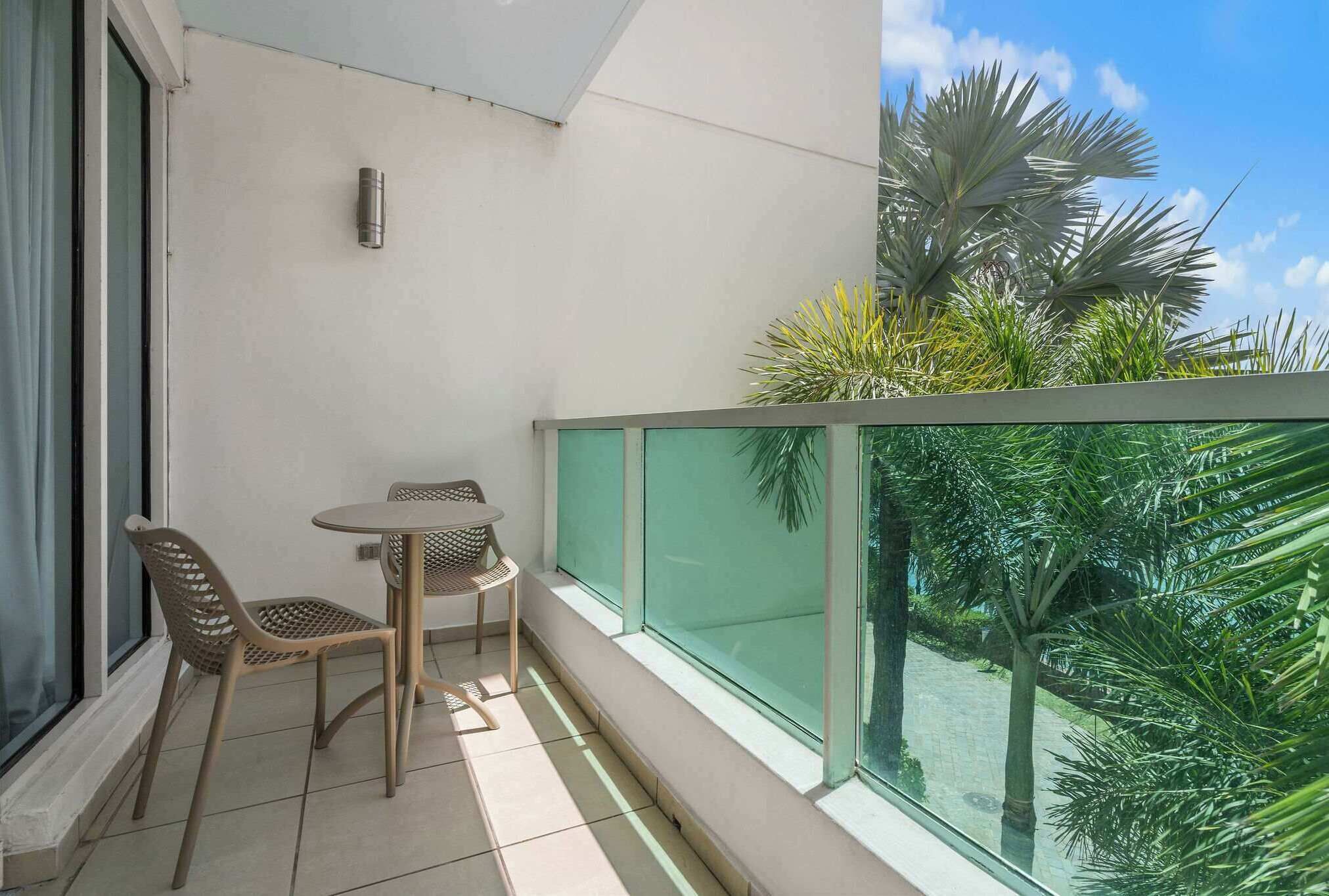 Un balcone con un tavolino e due sedie, ringhiera in vetro e vista su palme.