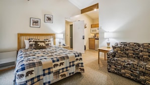 Ein Schlafzimmer mit einem großen Bett, einem Holzkopfteil, einer geblümten Steudecke, einem Nachttisch mit einer Lampe, einem Sessel mit einem Tierdruck und einer durchgehenden Tür erkennbaren Küche.