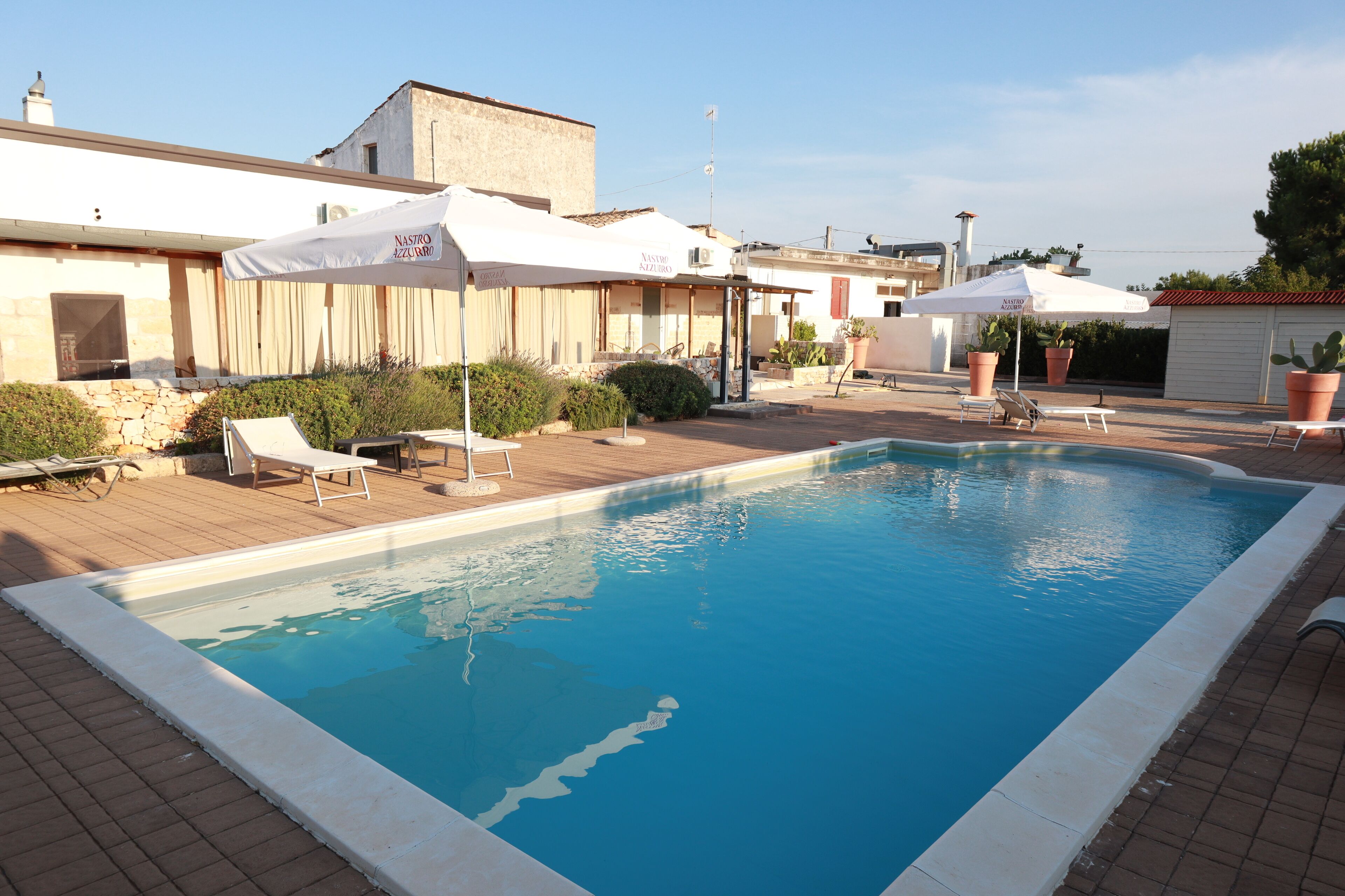 A swimming pool with lounge chairs and umbrellas, surrounded by buildings and potted plants.