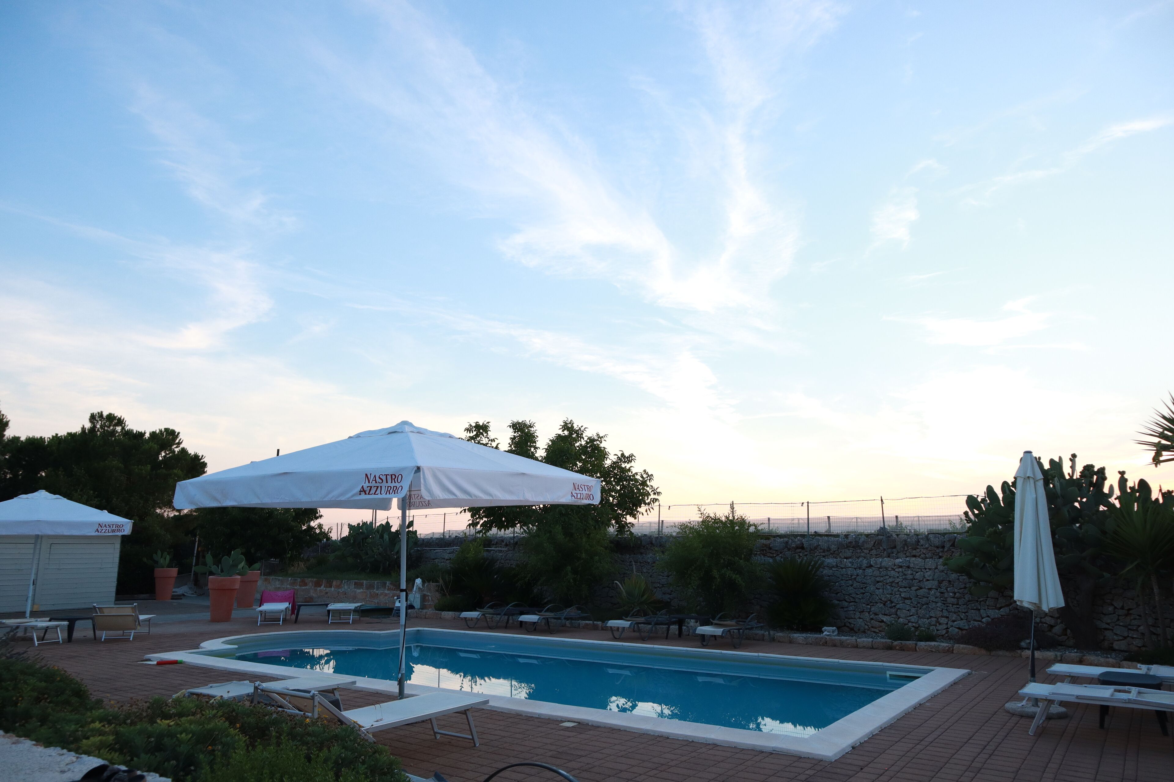 Ein Schwimmbecken mit Liegestühlen und Sonnenschirmen, umgeben von einer Steinmauer und viel Grün.