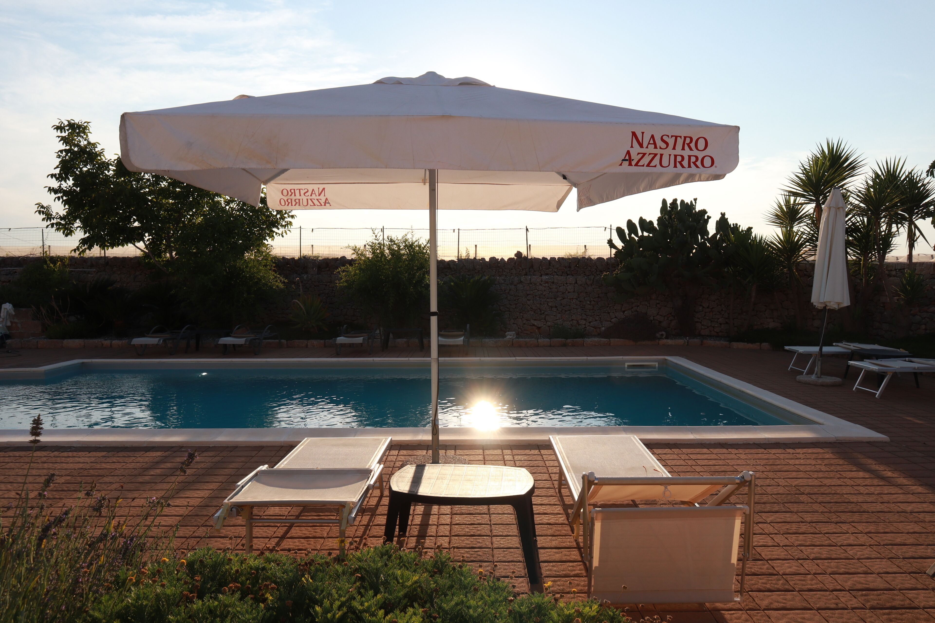 A poolside area with lounge chairs, a table, and a large umbrella labeled "Nastro Azzurro".