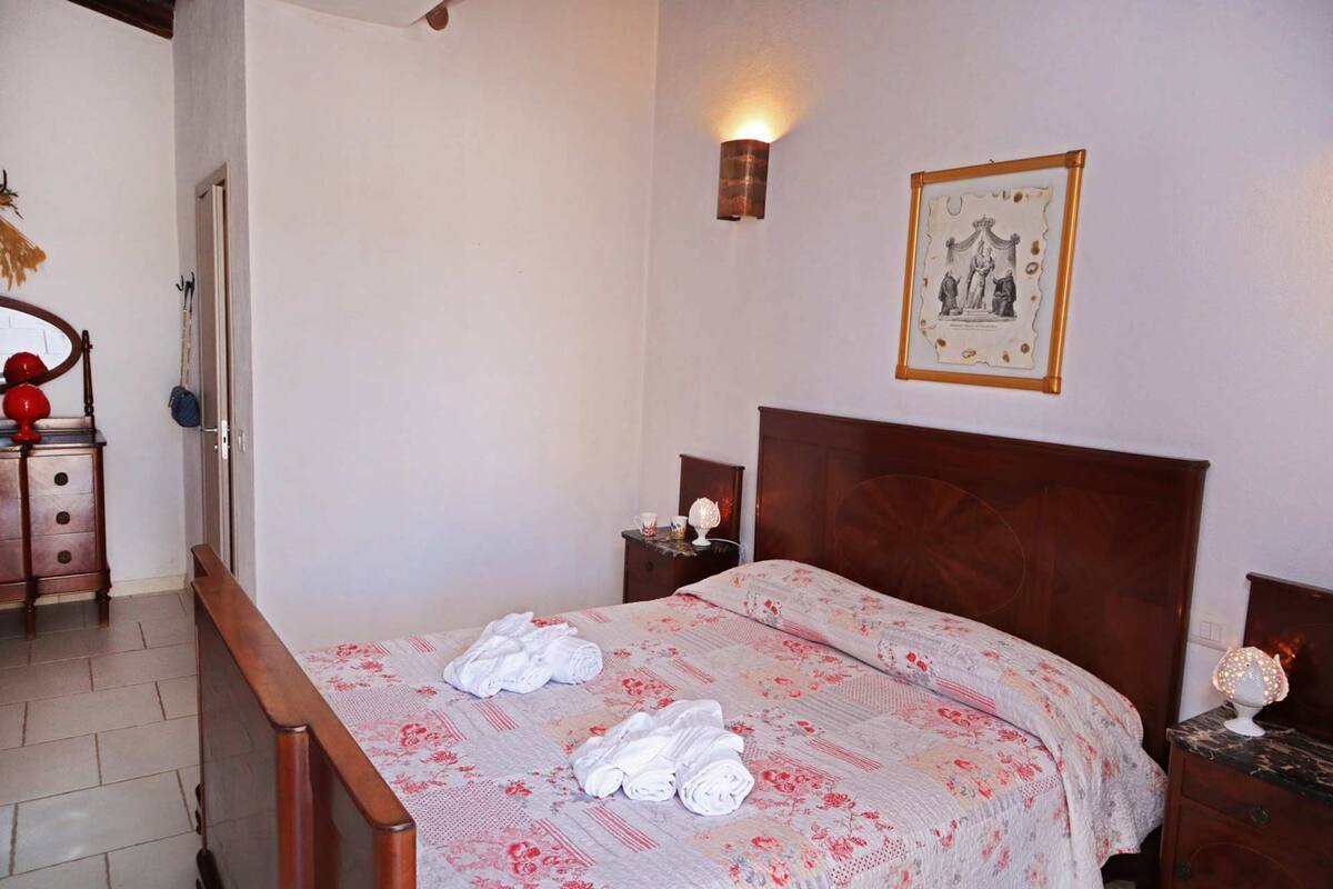 A neatly made bed with a floral bedspread and towels, a wooden headboard, a wall-mounted light, and a framed picture above the bed.