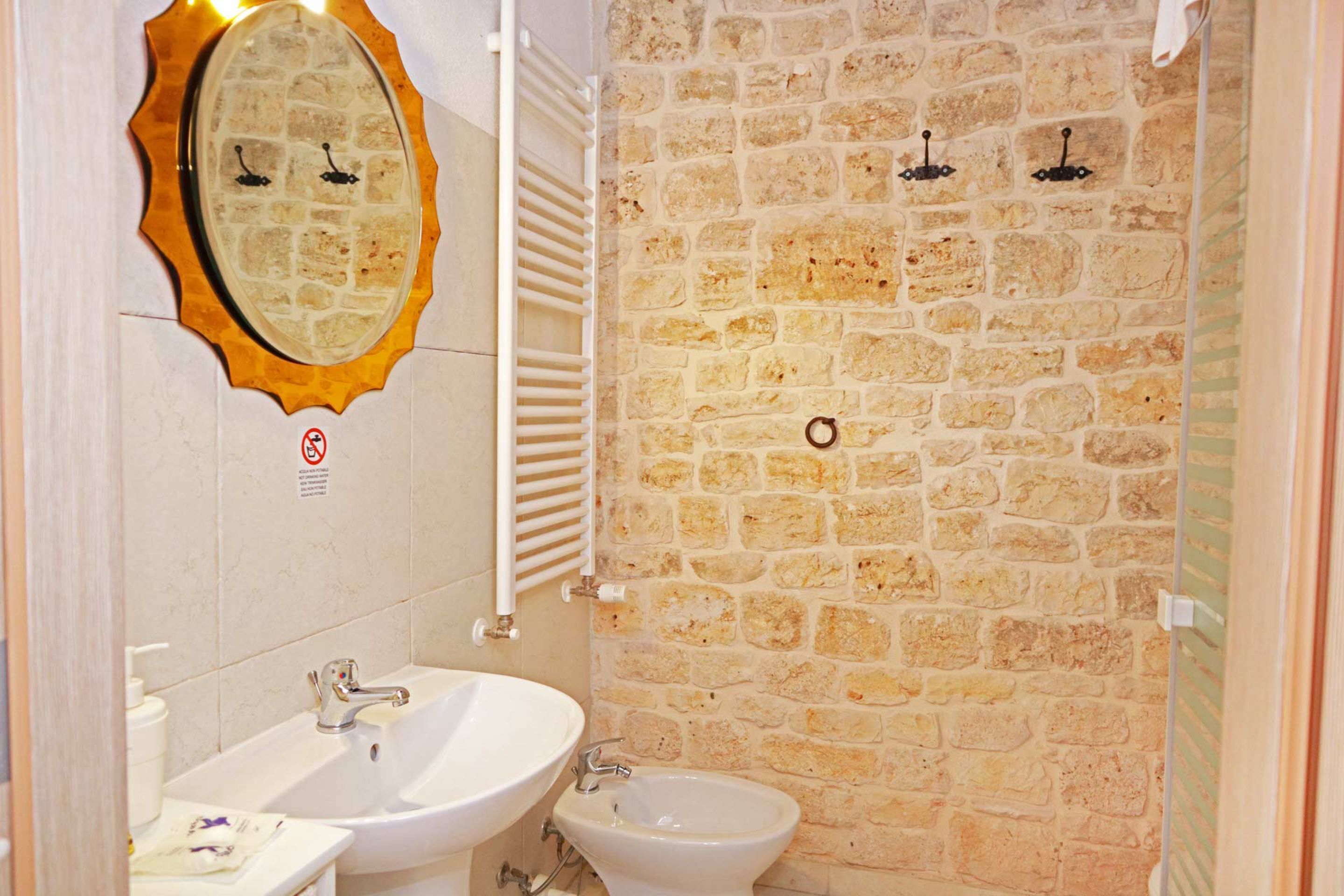 Un baño con pared de piedra, un lavabo blanco, un bidé y un espejo con marco dorado.
