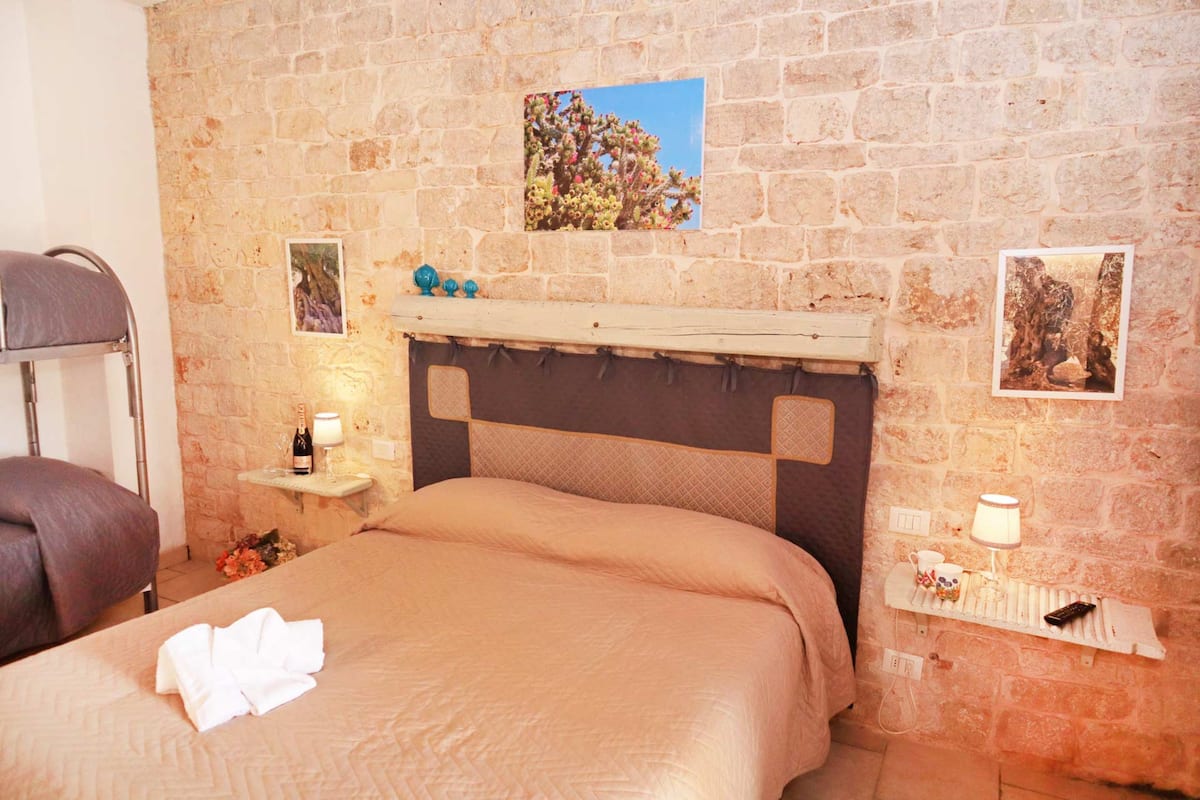 A bedroom with a stone wall, a bed with a headboard, a nightstand with a lamp, and a picture of a tree.