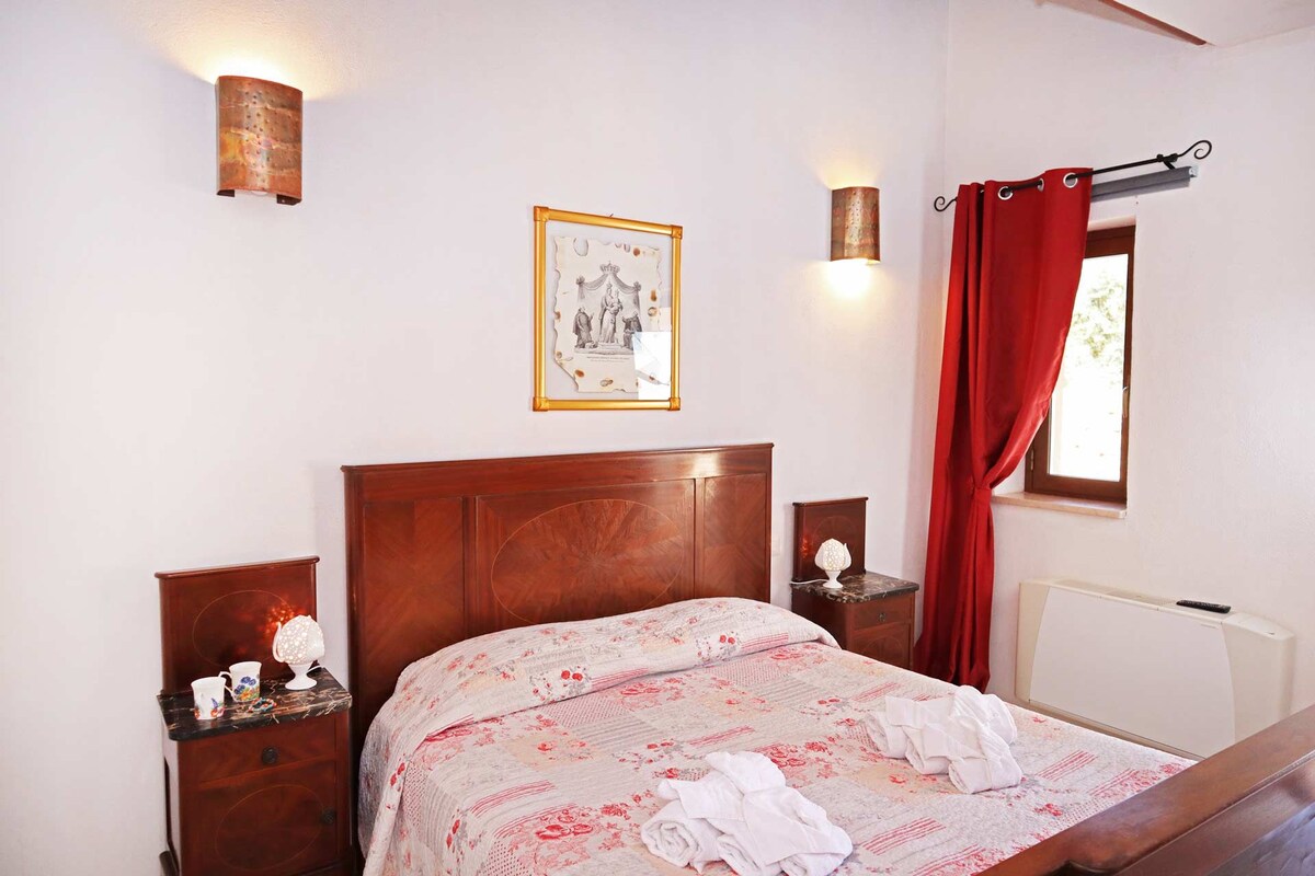 A neatly made bed with a floral bedspread, a framed picture on the wall, and a window with red curtains.