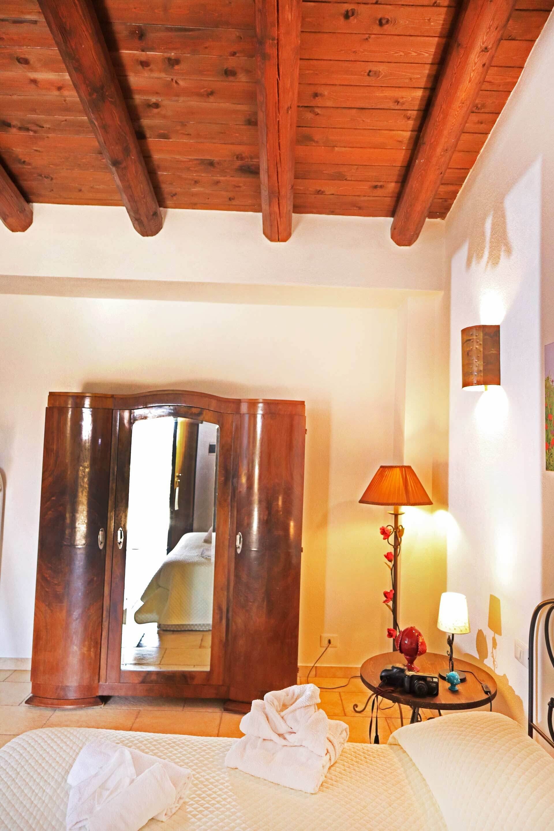 Una camera da letto con soffitto in legno, un letto con piumone bianco, un armadio in legno, un tavolino con lampada e uno specchio.